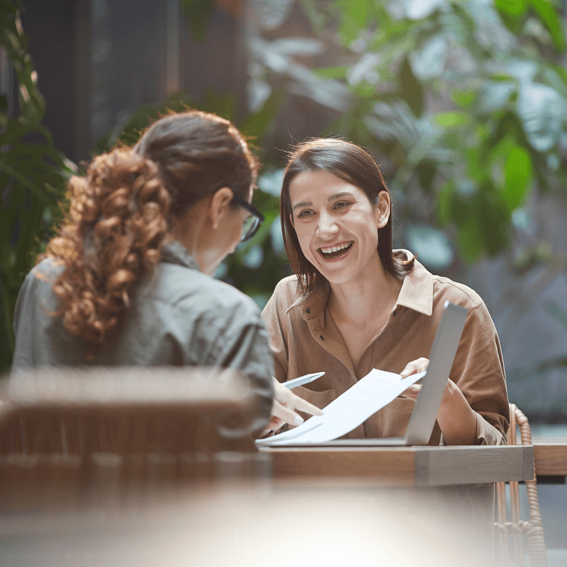 Two women smiling and discussing documents during a meeting in a bright, plant-filled workspace